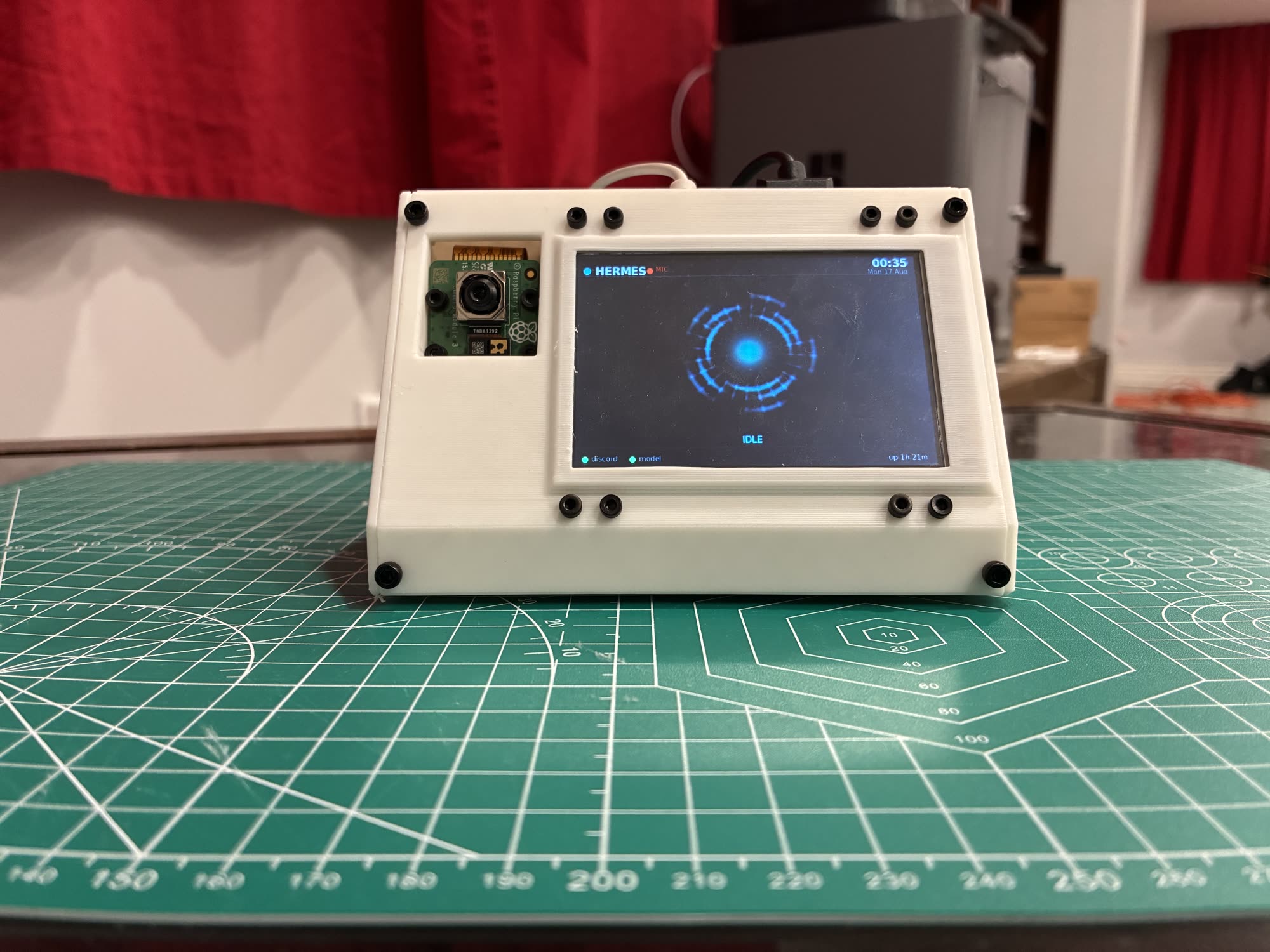 The finished Hermes Pi on a cutting mat: a white 3D-printed wedge enclosure with a Raspberry Pi camera module beside a lit panel reading HERMES and IDLE.