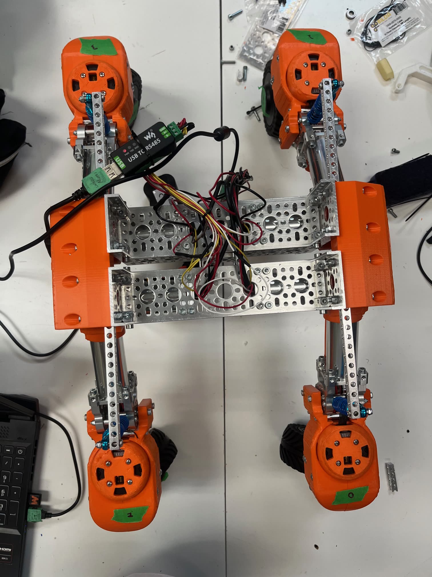 The swerve chassis from above: four orange 3D-printed swerve modules with coilover springs, linked by an aluminium frame with wiring routed through the centre.