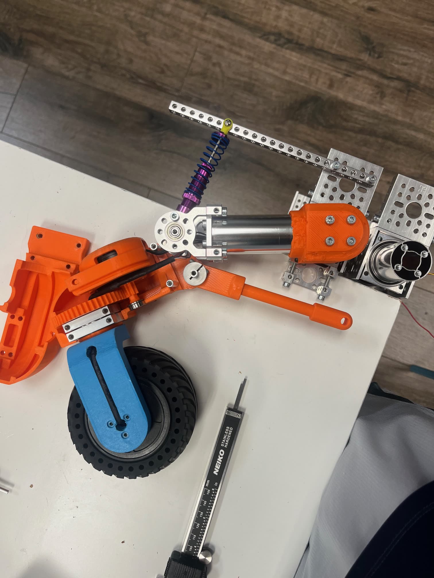 A single swerve module disassembled on a bench: the orange 3D-printed steering housing with its ring gear, a coilover spring and damper, the drive linkage, and the wheel with its blue mounting fork, beside a steel rule.